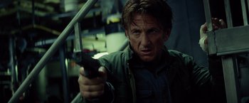 Movie still from “The Gunman” (2015), directed by Pierre Morel – A man holding a gun in a dark room; Close Up shot, Low angle