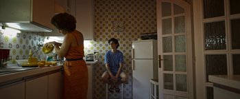 Movie still from “The Hand of God” (2021), directed by Paolo Sorrentino – A woman sitting on top of a stool in a kitchen; Wide shot, High angle