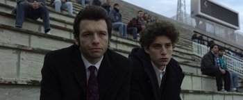 Movie still from “The Hand of God” (2021), directed by Paolo Sorrentino – Two men in suits and ties sitting next to each other in front of a group of people; Medium shot, Low angle