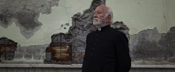 Movie still from “The Hand of God” (2021), directed by Paolo Sorrentino – An older man wearing a priest's outfit stands in front of a wall; Medium shot, Low angle