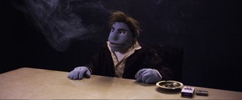 Movie still from “The Happytime Murders” (2018), directed by Brian Henson – A person wearing a puppet sitting at a table with smoke coming out of their mouth; Medium shot, Low angle