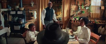Movie still from “The Harder They Fall” (2021), directed by Jeymes Samuel – A man standing in front of a table with a hat on it; Medium shot, Over the shoulder angle