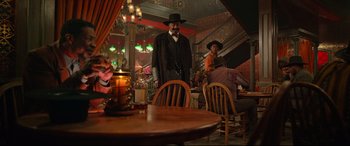 Movie still from “The Harder They Fall” (2021), directed by Jeymes Samuel – A man standing in front of a table in a room; Wide shot, Over the shoulder angle