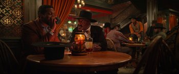 Movie still from “The Harder They Fall” (2021), directed by Jeymes Samuel – A man sitting at a table with a candle in front of him; Medium shot, Over the shoulder angle