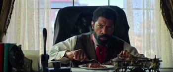 Movie still from “The Harder They Fall” (2021), directed by Jeymes Samuel – A man sitting at a table eating a piece of pizza; Close Up shot, Over the shoulder angle