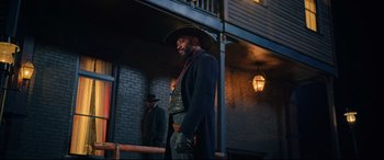 Movie still from “The Harder They Fall” (2021), directed by Jeymes Samuel – A man in a cowboy hat standing in front of a brick building; Medium shot, Over the shoulder angle