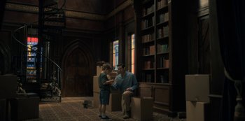 Movie still from “The Haunting of Hill House” (2018), directed by Mike Flanagan – A man and a boy sitting in a room with boxes in front of a bookcase; Wide shot, High angle