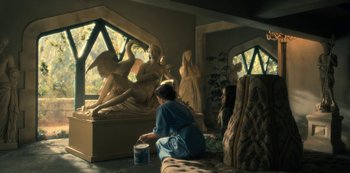 Movie still from “The Haunting of Hill House” (2018), directed by Mike Flanagan – A woman sitting on a couch in front of a statue; Wide shot, High angle