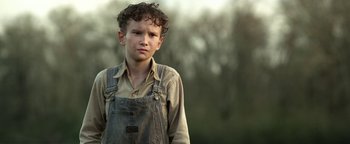 Movie still from “The Highwaymen” (2019), directed by John Lee Hancock – A young boy wearing overalls standing in front of trees; Close Up shot, Low angle