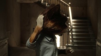 Movie still from “The House” (2022), directed by Paloma Baeza – A person with a cat's head in a room with stairs; Close Up shot, Over the shoulder angle