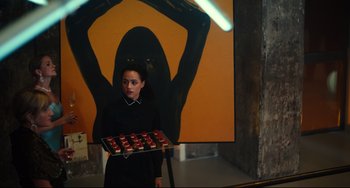 Movie still from “The Invitation” (2022), directed by Jessica M. Thompson – A woman holding a tray of donuts in front of a painting; Medium shot, Low angle