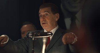 Movie still from “The Irishman” (2019), directed by Martin Scorsese – A man in a gray suit and red neck tie speaking at a microphone; Close Up shot, Low angle