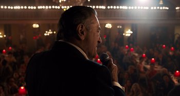 Movie still from “The Irishman” (2019), directed by Martin Scorsese – An older man is holding a microphone in front of an audience; Medium shot, Over the shoulder angle