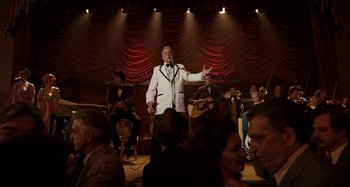 Movie still from “The Irishman” (2019), directed by Martin Scorsese – A group of people sitting around a stage with a man in a white suit; Wide shot, Over the shoulder angle