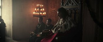 Movie still from “The King” (2019), directed by David Michôd – A group of people sitting in a room with candles in the background; Medium shot, Low angle