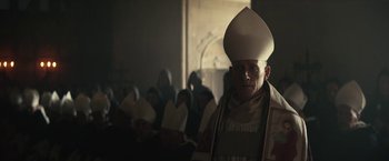 Movie still from “The King” (2019), directed by David Michôd – A man wearing a priest's outfit and a white hat; Medium shot, Low angle