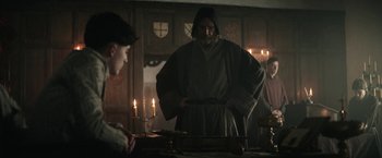 Movie still from “The King” (2019), directed by David Michôd – A man in a hooded robe stands in front of a table with candles; Medium shot, Over the shoulder angle