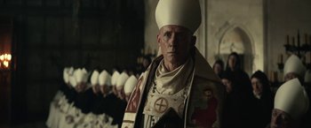 Movie still from “The King” (2019), directed by David Michôd – A man wearing a priest's outfit in front of a group of people; Close Up shot, Over the shoulder angle