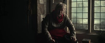 Movie still from “The King” (2019), directed by David Michôd – An older man sitting in a chair in a room; Medium shot, Over the shoulder angle