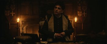 Movie still from “The King” (2019), directed by David Michôd – A man sitting at a table in a dark room; Medium shot, Low angle