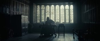 Movie still from “The King” (2019), directed by David Michôd – A person sitting at a desk in front of a window; Wide shot, Low angle