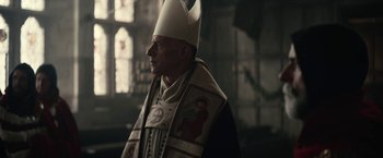 Movie still from “The King” (2019), directed by David Michôd – A man wearing a priest's hat and a priest's robe; Medium shot, Low angle