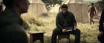 Movie still from “The King” (2019), directed by David Michôd – A man sitting on top of a wooden bench in front of tents; Medium shot, Low angle