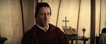 Movie still from “The King” (2019), directed by David Michôd – A man wearing a monk's outfit with a cross in the background; Close Up shot, Over the shoulder angle