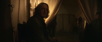 Movie still from “The King” (2019), directed by David Michôd – A man with long white hair and glasses is sitting in a dimly - lit room; Medium shot, Over the shoulder angle