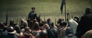 Movie still from “The King” (2019), directed by David Michôd – A group of people in medieval garb gathered in a field; Wide shot, Over the shoulder angle