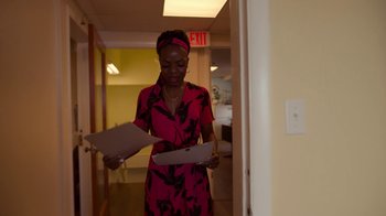 Movie still from “The Laundromat” (2019), directed by Steven Soderbergh – A woman in a pink dress holding papers in her hands; Medium shot, Over the shoulder angle