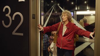 Movie still from “The Laundromat” (2019), directed by Steven Soderbergh – An older woman in a red jacket standing in front of a door; Medium shot, Over the shoulder angle