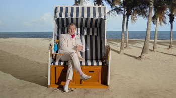 Movie still from “The Laundromat” (2019), directed by Steven Soderbergh – A man sitting on top of a chair on the beach; Wide shot, Low angle