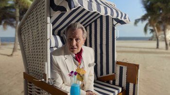 Movie still from “The Laundromat” (2019), directed by Steven Soderbergh – An older man sitting under a canopy with a drink in his hand; Medium shot, Over the shoulder angle