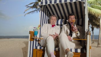 Movie still from “The Laundromat” (2019), directed by Steven Soderbergh – A couple of men sitting on top of a beach chair; Medium shot, Low angle