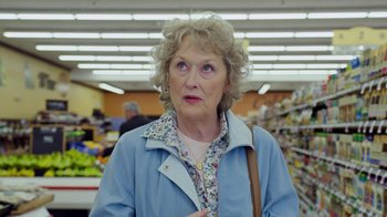 Movie still from “The Laundromat” (2019), directed by Steven Soderbergh – An older woman in a blue jacket is standing in a grocery store; Close Up shot, Over the shoulder angle