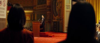 Movie still from “The Laundromat” (2019), directed by Steven Soderbergh – A man standing at a podium in front of an audience; Extreme Wide shot, High angle