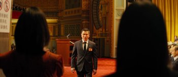 Movie still from “The Laundromat” (2019), directed by Steven Soderbergh – A man in a suit and tie standing in front of an audience; Medium shot, Over the shoulder angle