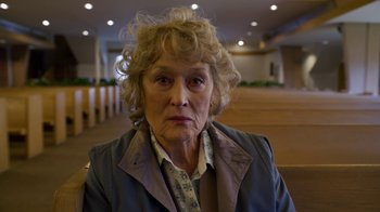 Movie still from “The Laundromat” (2019), directed by Steven Soderbergh – An older woman with blonde curly hair sitting in front of a wooden bench; Close Up shot, High angle