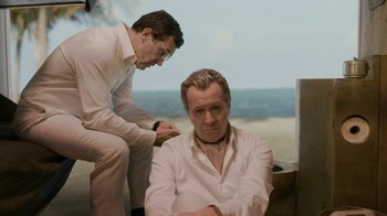Movie still from “The Laundromat” (2019), directed by Steven Soderbergh – A man sitting next to another man on a beach; Medium shot, High angle