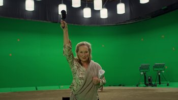 Movie still from “The Laundromat” (2019), directed by Steven Soderbergh – A woman holding up a microphone in front of a green screen; Medium shot, High angle