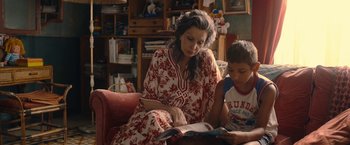 Movie still from “The Life Ahead” (2020), directed by Edoardo Ponti – A woman sitting on a couch reading a book to a child; Medium shot, Over the shoulder angle