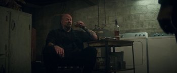 Movie still from “The Man from Toronto” (2022), directed by Patrick Hughes – A man sitting at a table with a drink in his hand; Medium shot, Over the shoulder angle