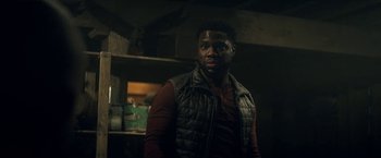 Movie still from “The Man from Toronto” (2022), directed by Patrick Hughes – A man standing in a dark room wearing a vest; Medium shot, Over the shoulder angle