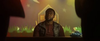 Movie still from “The Man from Toronto” (2022), directed by Patrick Hughes – A man sitting at a table in front of a house; Medium shot, Over the shoulder angle