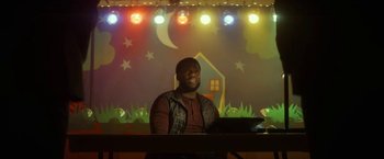 Movie still from “The Man from Toronto” (2022), directed by Patrick Hughes – A man sitting at a table in front of lights; Medium shot, Low angle