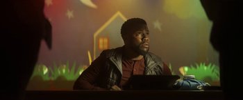 Movie still from “The Man from Toronto” (2022), directed by Patrick Hughes – A man sitting at a table in front of a projector screen; Medium shot, Low angle