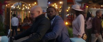 Movie still from “The Man from Toronto” (2022), directed by Patrick Hughes – A man leaning on another man's shoulder in front of lights; Medium shot, Over the shoulder angle
