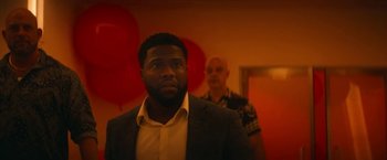Movie still from “The Man from Toronto” (2022), directed by Patrick Hughes – A man standing next to another man in a room with red balloons; Medium shot, Over the shoulder angle