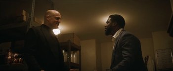 Movie still from “The Man from Toronto” (2022), directed by Patrick Hughes – Two men in suits talking to each other in a dimly - lit room; Medium shot, Over the shoulder angle